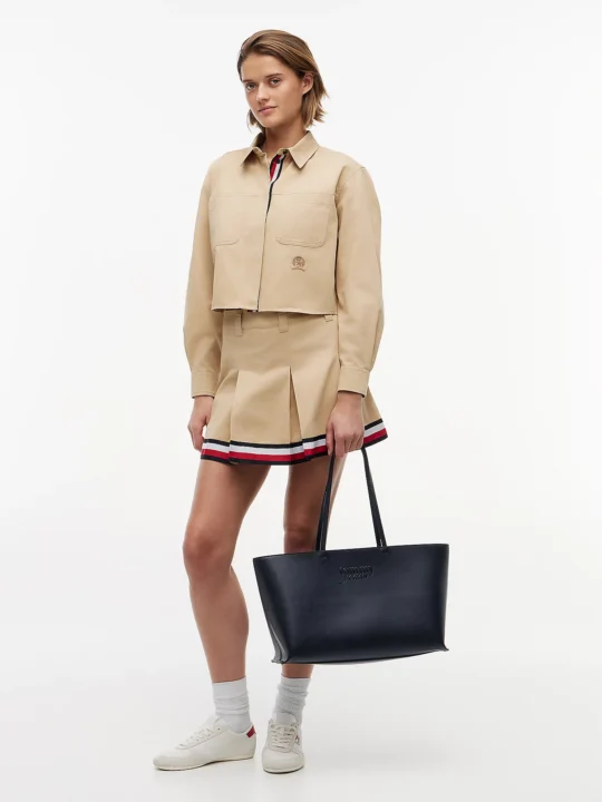 Model wearing a beige cropped utility jacket with two front pockets and a matching pleated mini skirt, holding a black tote bag.