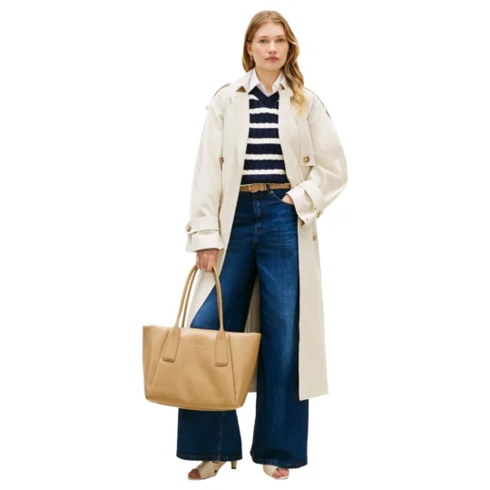 Person in a beige trench coat over a navy striped sweater and blue jeans, carrying a tan tote bag and wearing beige sandals.