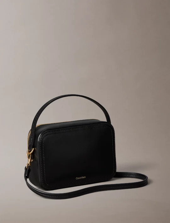 Black leather Calvin Klein handbag with gold zipper and strap, photographed against a neutral beige background, partial view shows the top and side of the bag.