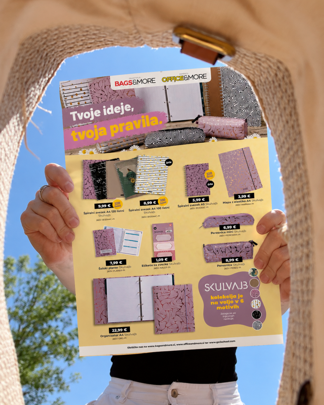 Hands holding up a yellow advertisement flyer for notebooks and planners with purple text, shown against a clear blue sky.